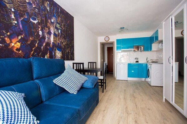 A modern living room with a blue sofa and a cityscape painting, a dining area with chairs, and a kitchen with blue cabinets and white appliances.