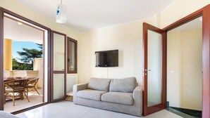A living room with a sofa, a flat-screen TV mounted on the wall, and a sliding glass door leading to an outdoor area with a dining table and chairs.