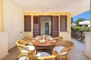 Un comedor con una mesa redonda dispuesta para seis personas, rodeada de sillas de mimbre, en un balcón con vistas a los edificios y la vegetación circundantes.