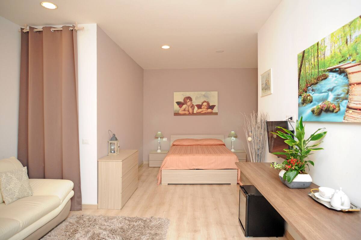 A modern bedroom with a bed, bedside tables, a desk, a lamp, a plant, and a painting on the wall.