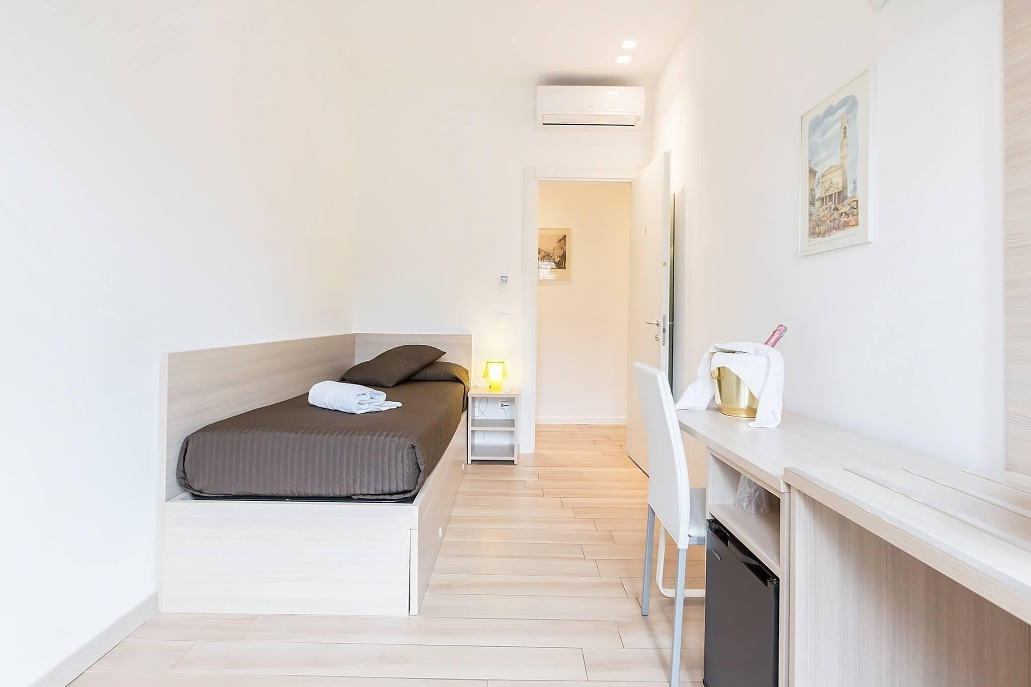 Quarto compacto com cama, mesa de cabeceira, abajur e pequena área de cozinha.