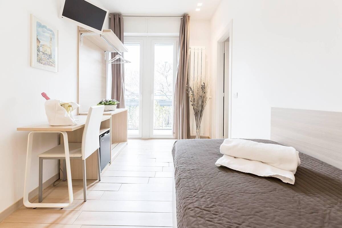 A modern hotel room with a bed, a desk, a chair, a television, and a window with curtains.