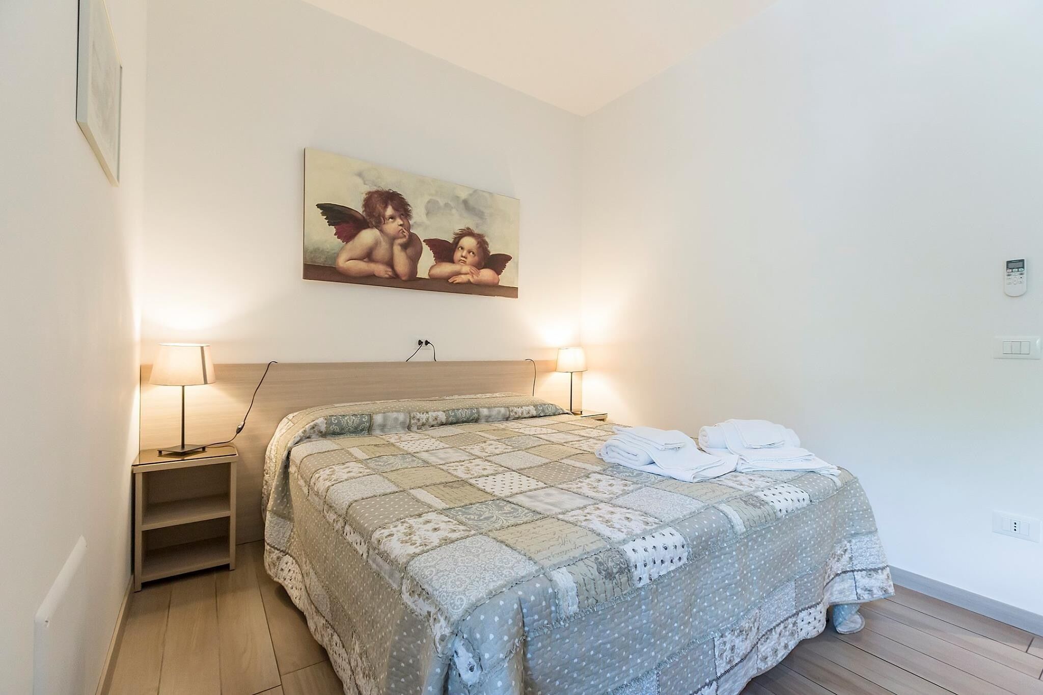 A bedroom with a bed, two bedside lamps, a nightstand, a painting of cherubs, and a white wall.