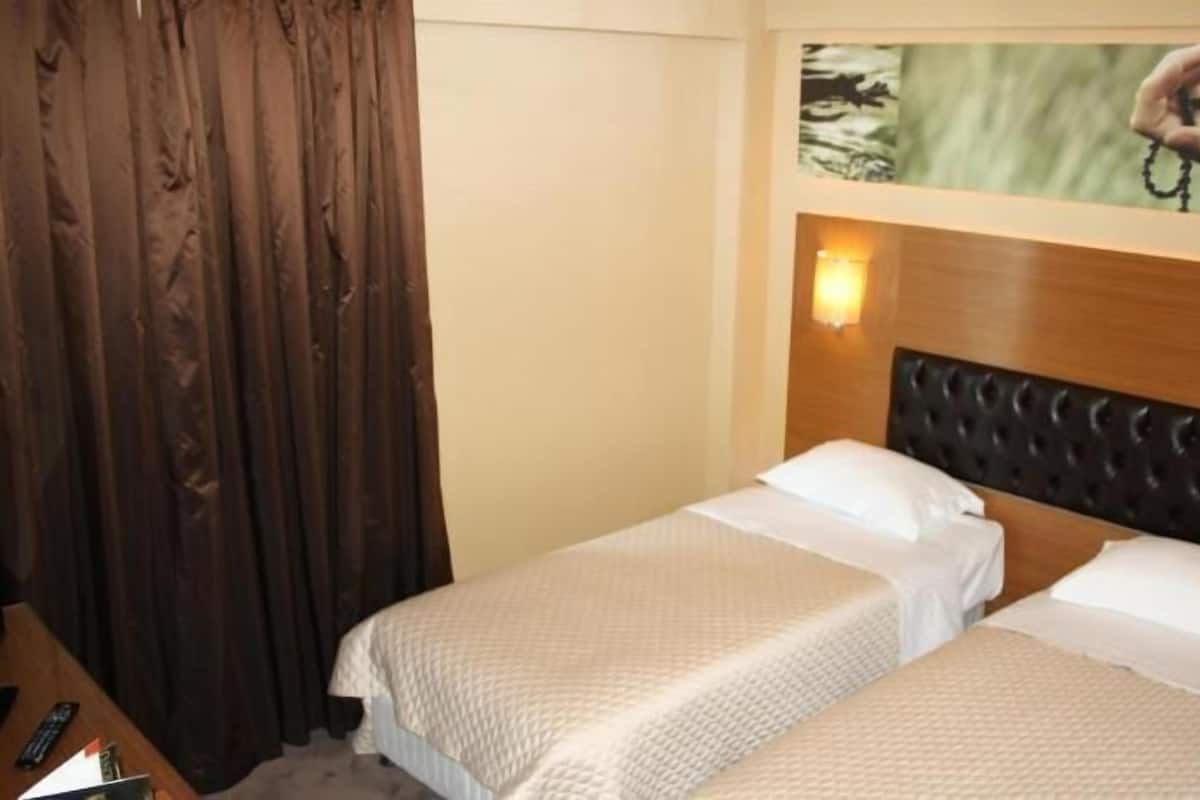 A hotel room with two single beds, a wooden headboard, a painting above the beds, and a curtain on the left.
