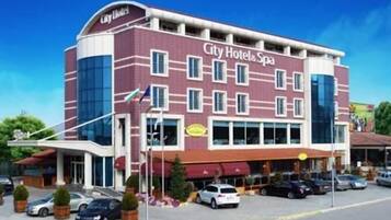 A City Hotel & Spa with a modern design, featuring a prominent red and white facade, multiple windows, and a parking area with several cars.