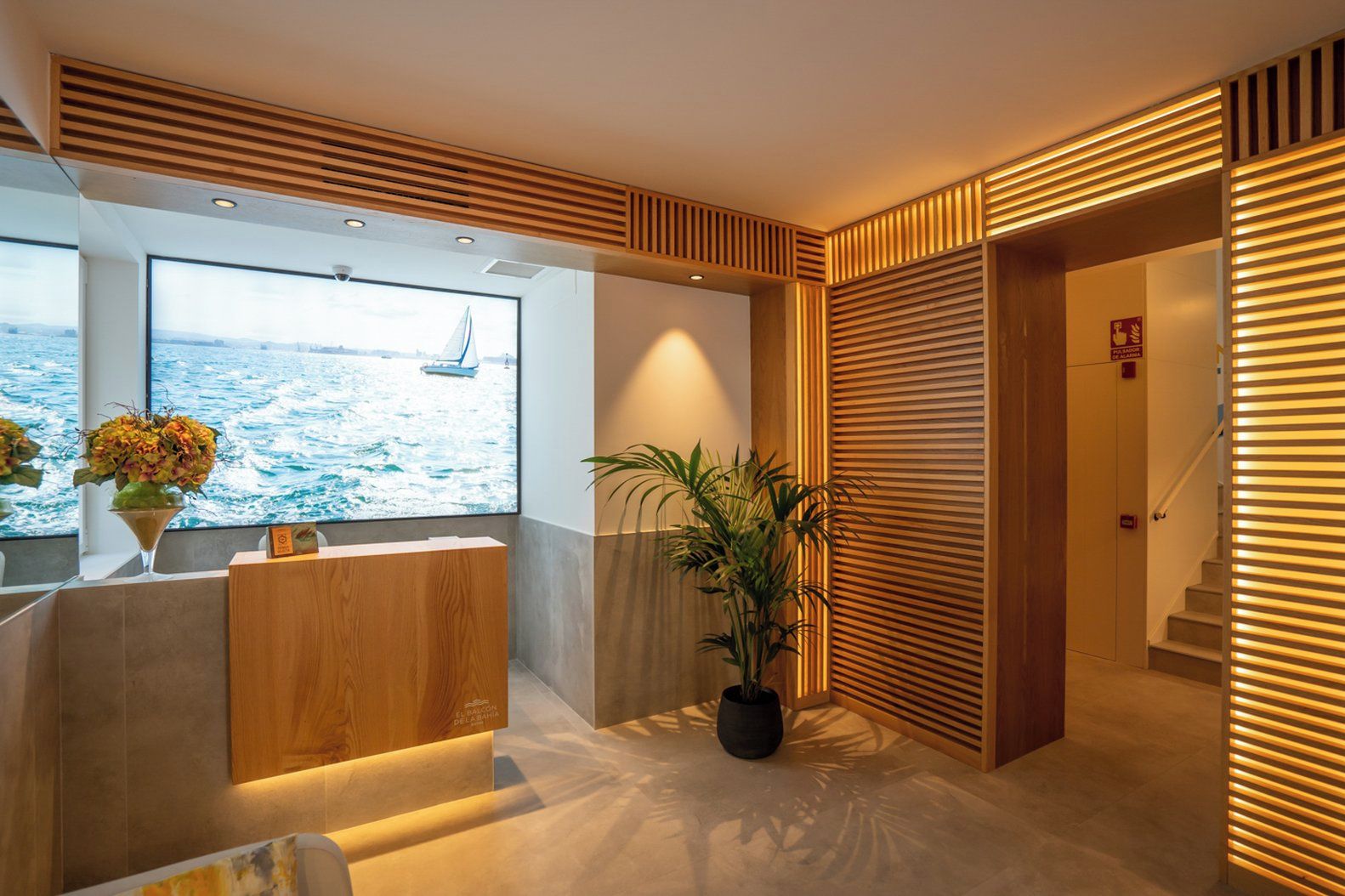 Una moderna zona de recepción con un amplio ventanal con vistas al mar, un mostrador de recepción, una planta en maceta y una escalera con barandillas iluminadas.