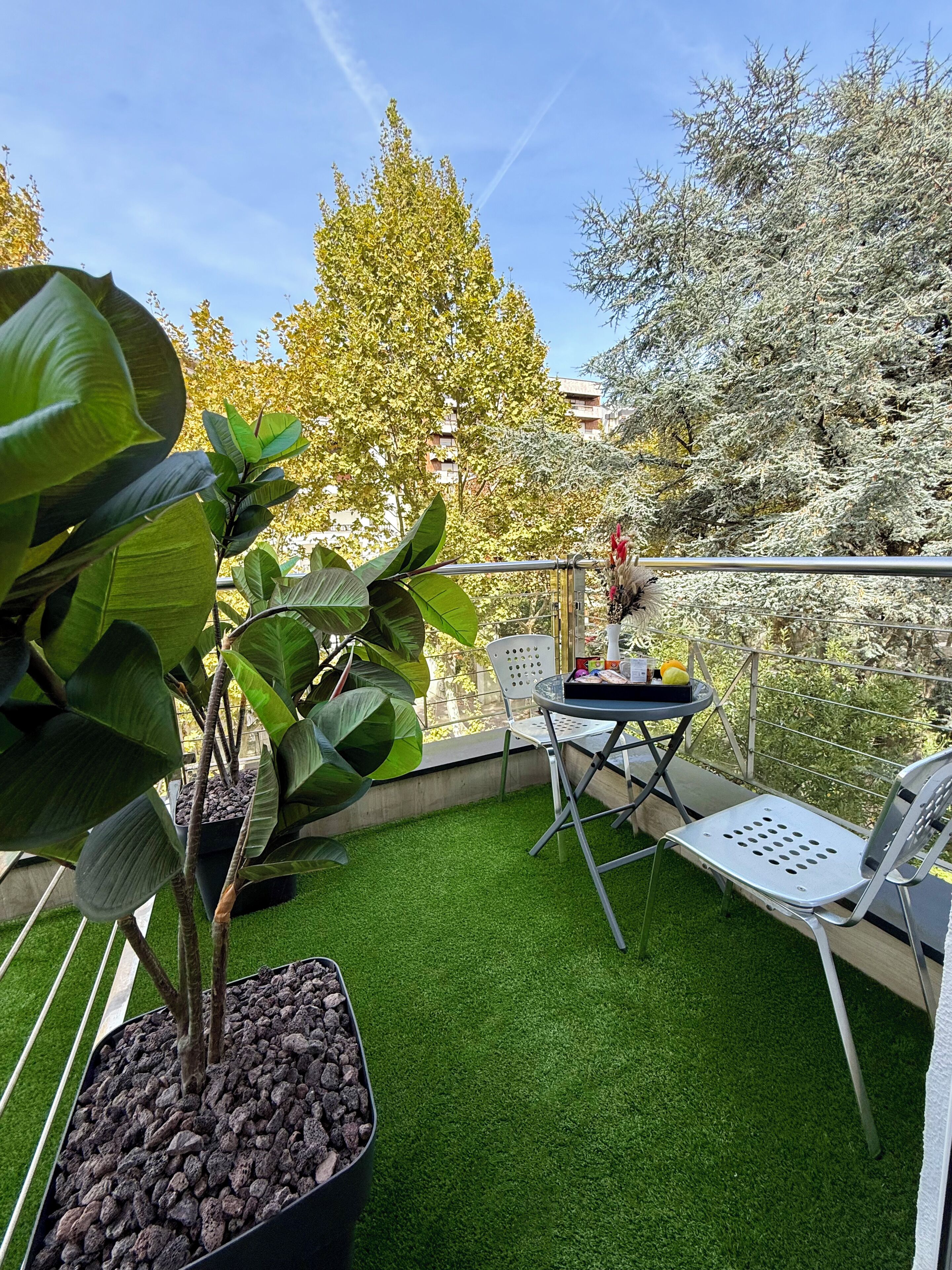 Un balcón con césped artificial, plantas en macetas, una mesa con sillas y vistas a árboles y edificios.