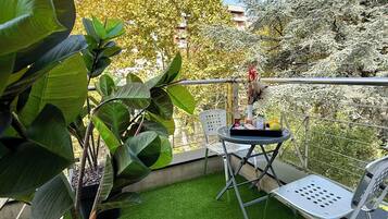 Un balcón con césped artificial, plantas en macetas, una mesa con sillas y vistas a árboles y edificios.