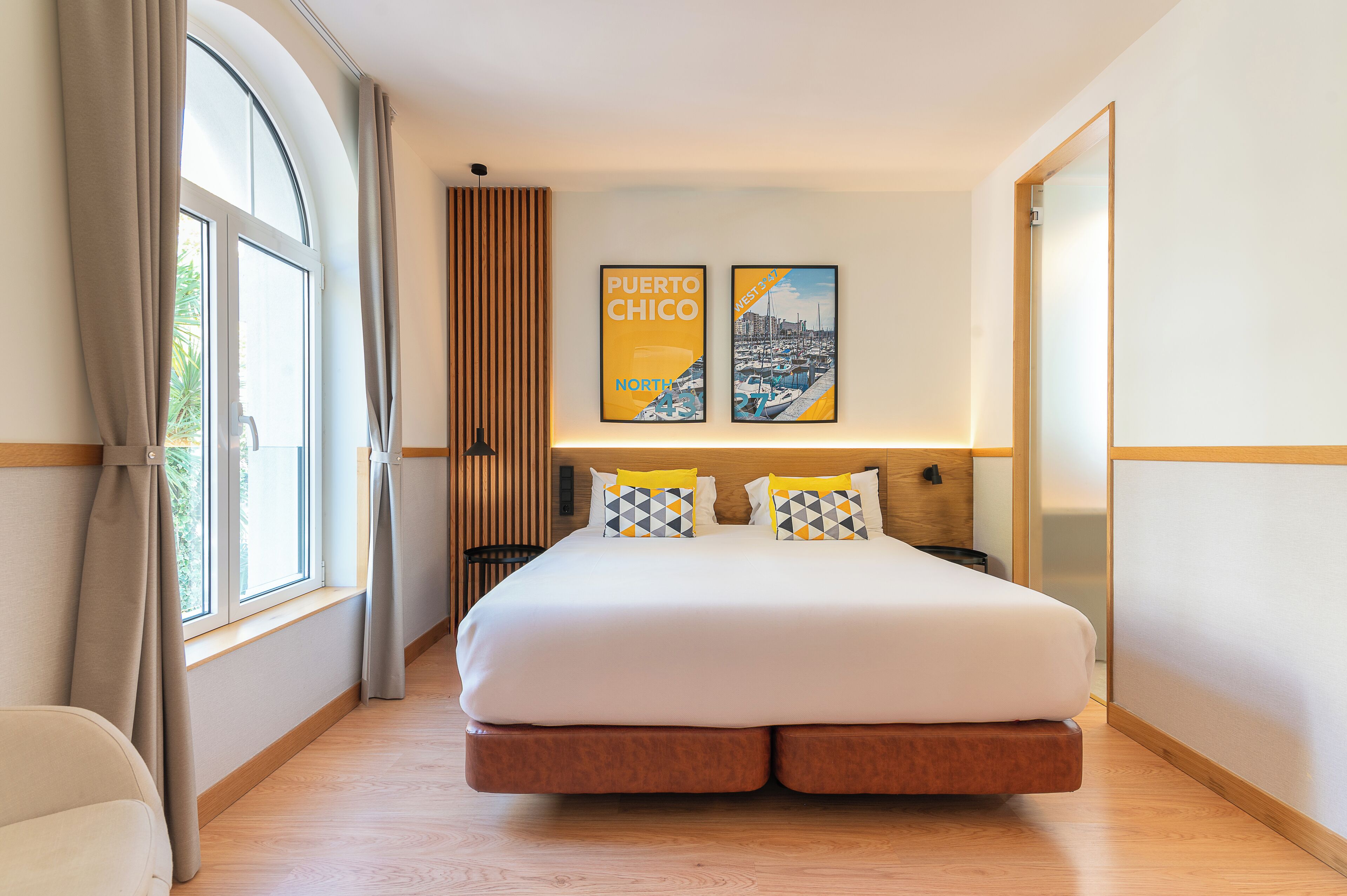 A modern hotel room with a large bed, a window with curtains, and two framed pictures on the wall.