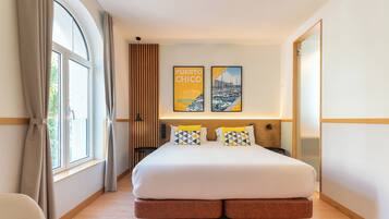 A modern hotel room with a large bed, a window with curtains, and two framed pictures on the wall.