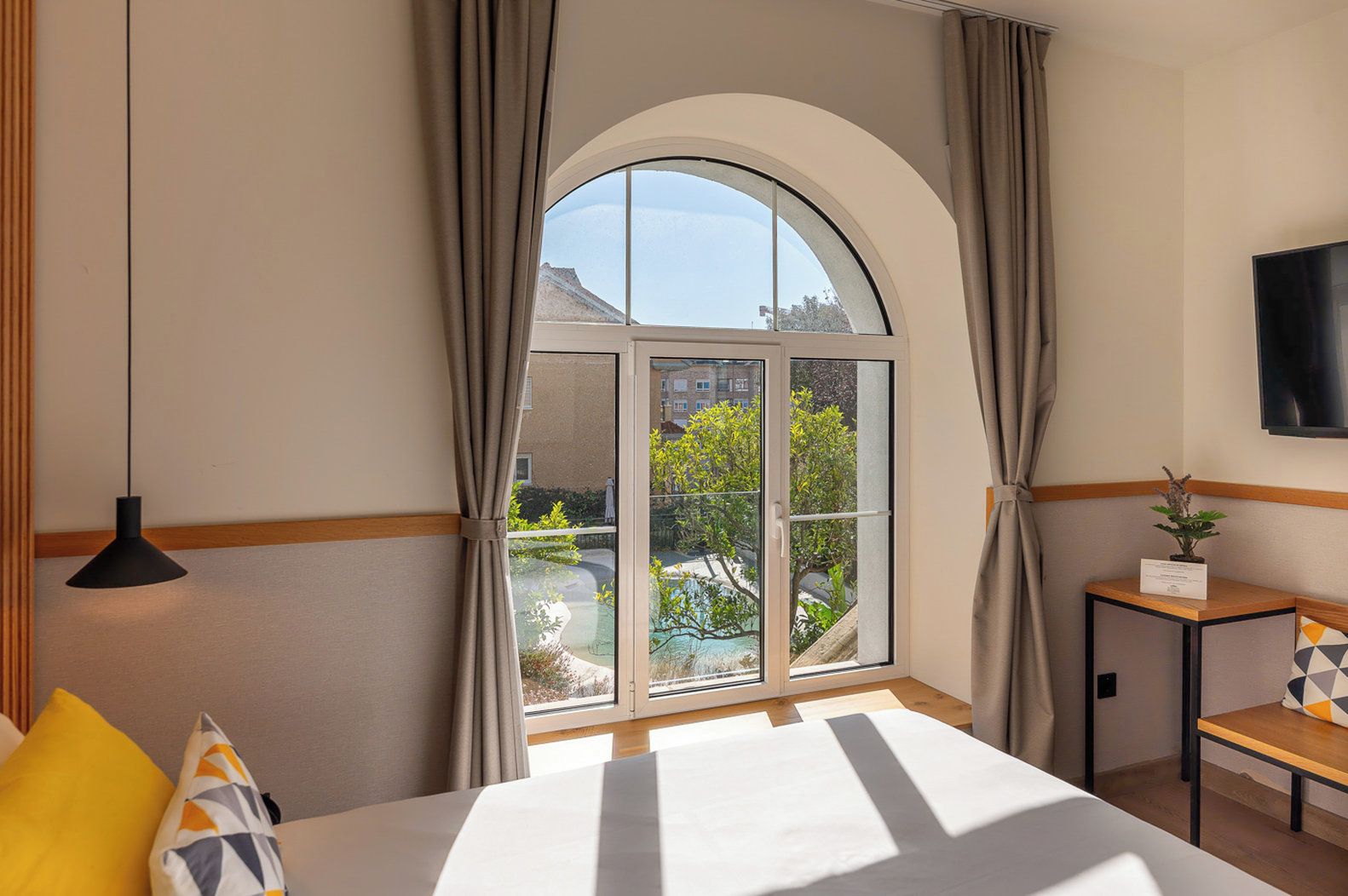 A hotel room with a large arched window, a bed, a bedside table with a plant, and a TV on the wall.