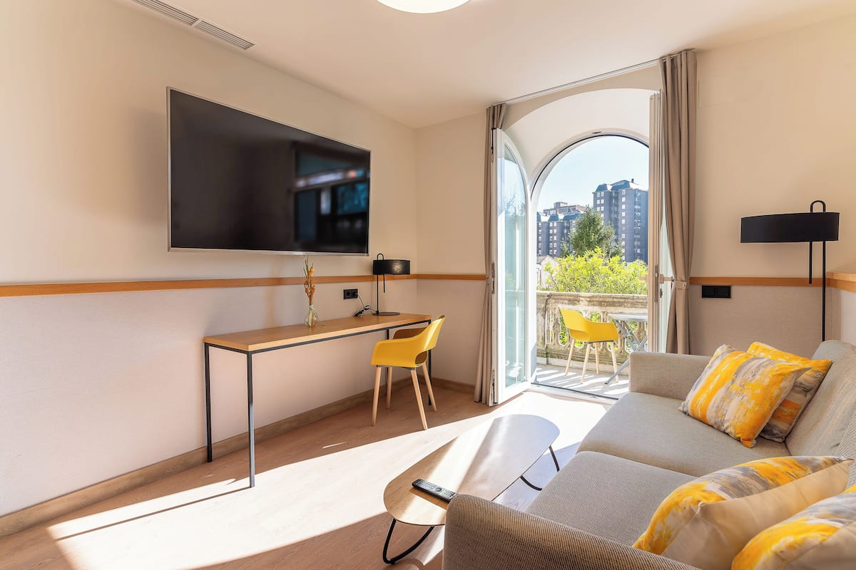 Ein modernes Hotelzimmer mit einem wandmontierten Flachbildfernseher, einem Holztisch mit Stuhl, einem Sofa mit gelben Kissen und einem großen Fenster mit Blick auf die Stadt.