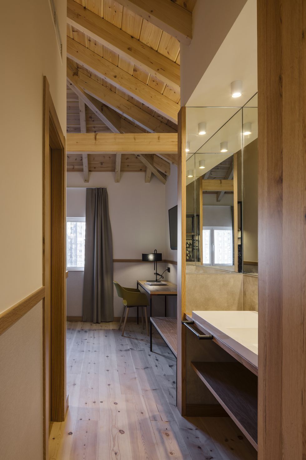 A compact room with a wooden ceiling, a desk with a chair, a bed, and a large mirror.