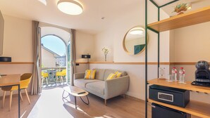 Una habitación de hotel moderna con sofá, una pequeña mesa de comedor, cafetera y un refrigerador pequeño.