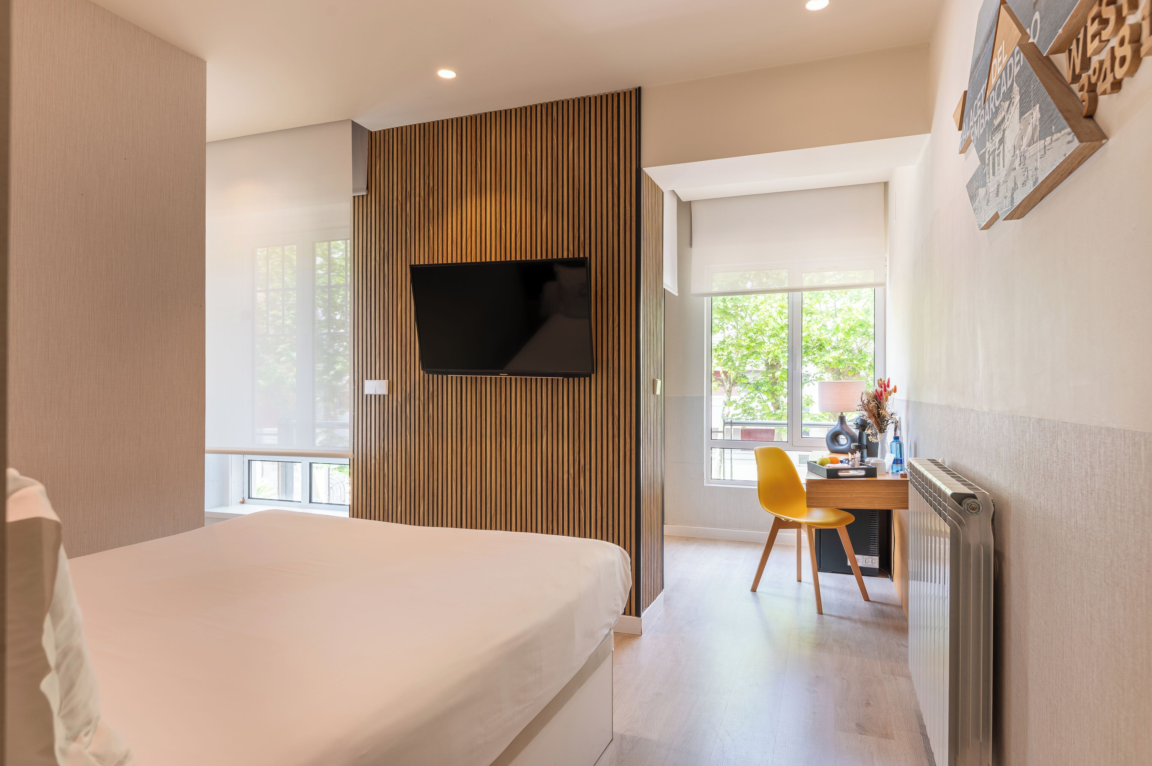 A modern hotel room with a bed, a desk with a chair, a flat-screen TV mounted on a wooden panel, and a window providing a view of greenery.