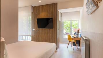 A modern hotel room with a bed, a desk with a chair, a flat-screen TV mounted on a wooden panel, and a window providing a view of greenery.