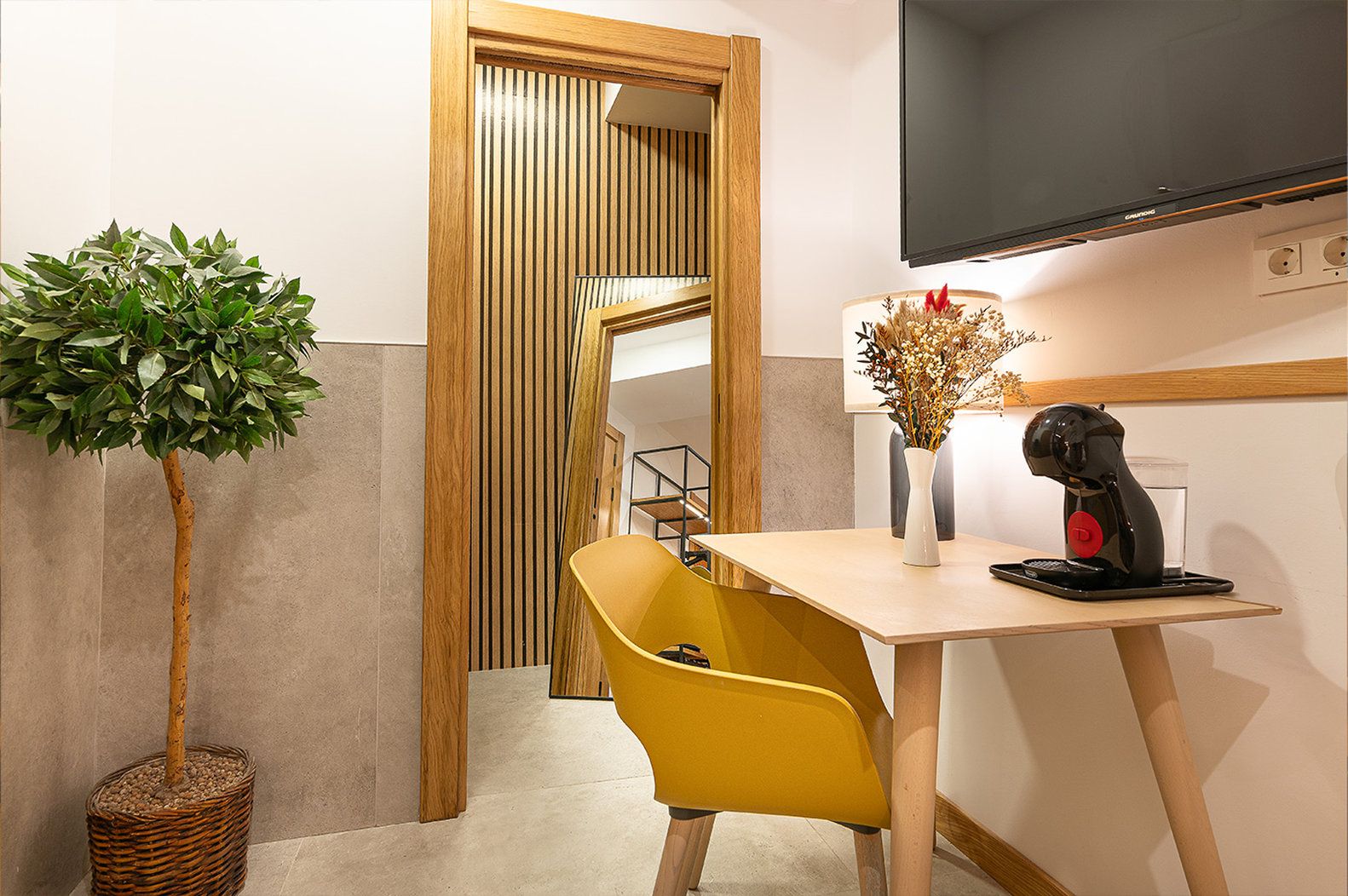 Una habitación moderna con una puerta de madera, una planta en maceta, un sillón amarillo, una mesa con un florero y una cafetera, y un televisor montado en la pared.