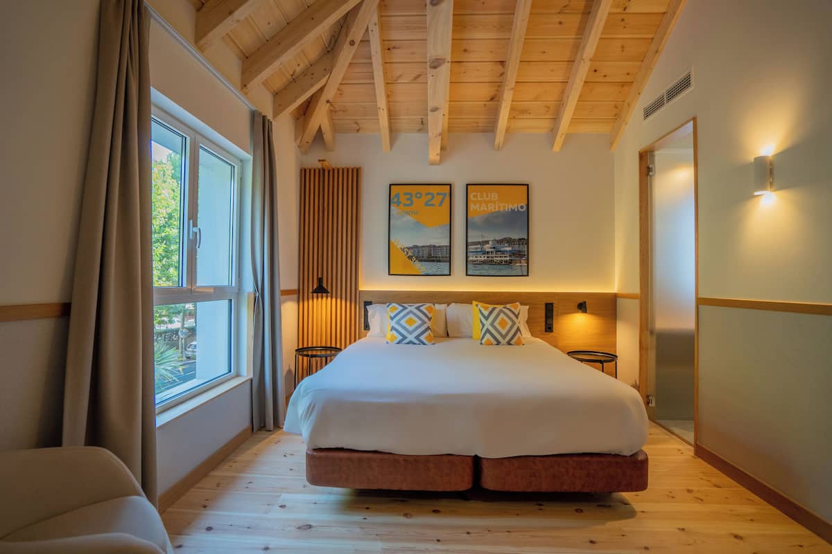 Ein Schlafzimmer mit einem großen Bett, Holzbalken an der Decke, einem Fenster mit Vorhängen und zwei Bildern an der Wand.