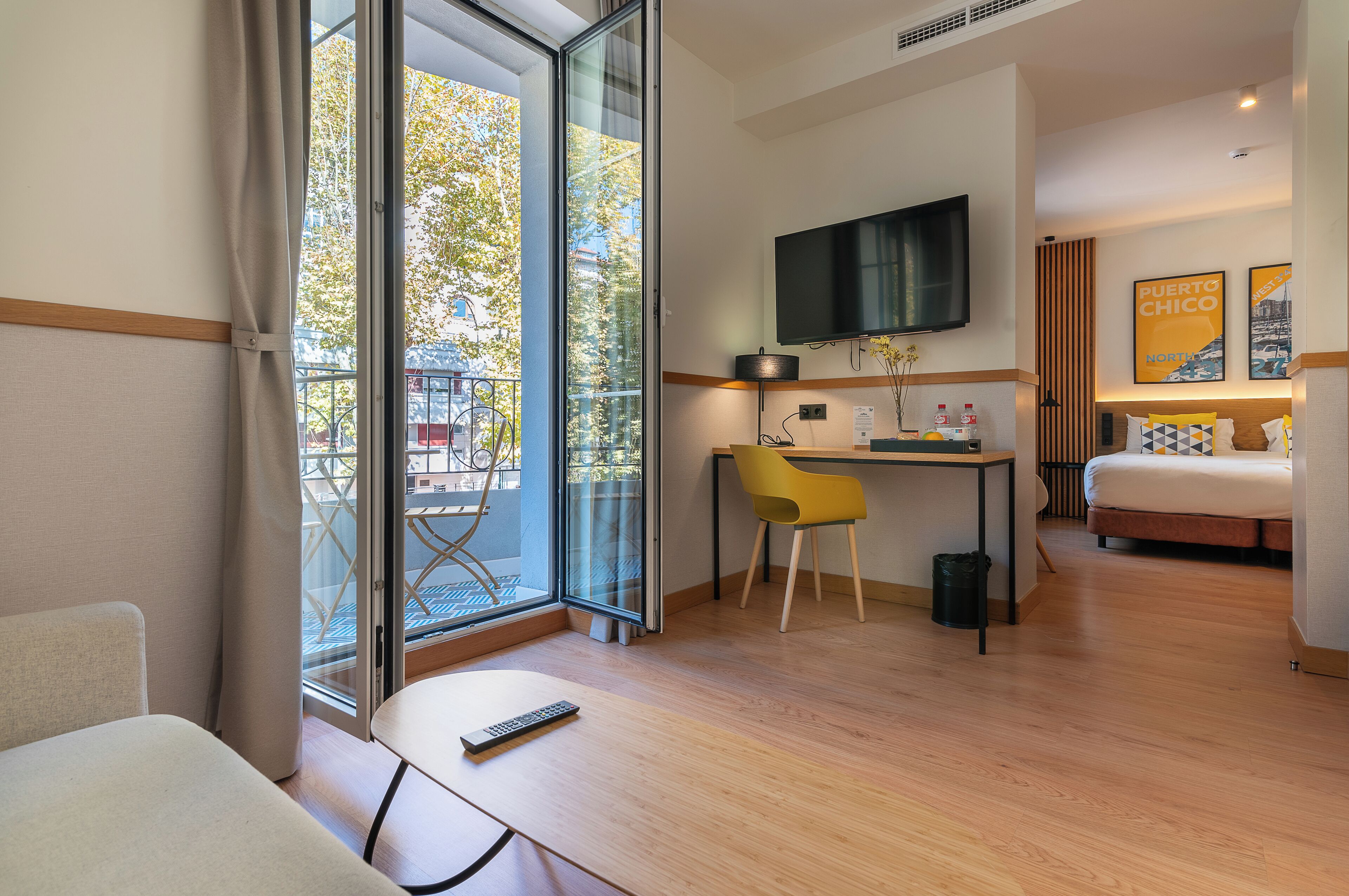 A hotel room with a bed, a desk with a chair, a TV mounted on the wall, and a balcony with a view of trees.