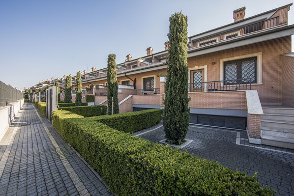 A row of modern brick buildings with balconies, a paved walkway, and neatly trimmed hedges. - Borgo Aureo (Rome)
