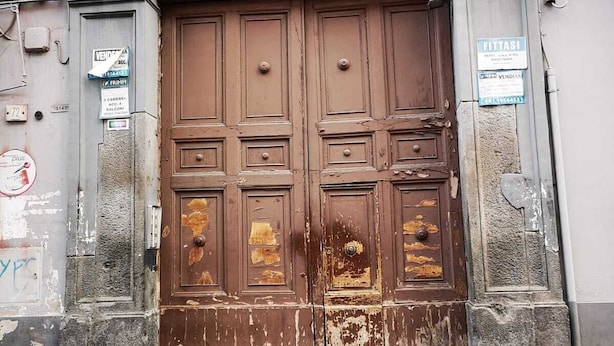 A large, ornate wooden door with a metal grille above it, set in a stone building with signs on the walls. - Dimora Ruggiero (Naples)
