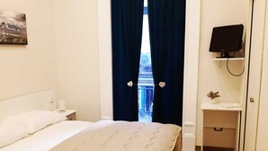 A hotel room with a bed, a bedside table, a TV, and a view of a building through a window with curtains. - Dimora Ruggiero (Naples)