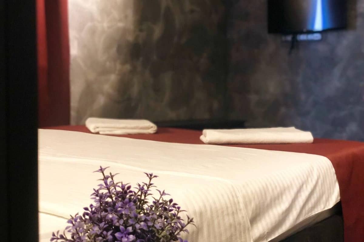 Ein Zimmer mit einem Bett, das mit weißen Leinen bezogen ist, einem wandmontierten Flachbildfernseher und einer Vase mit violetten Blumen auf dem Bett.