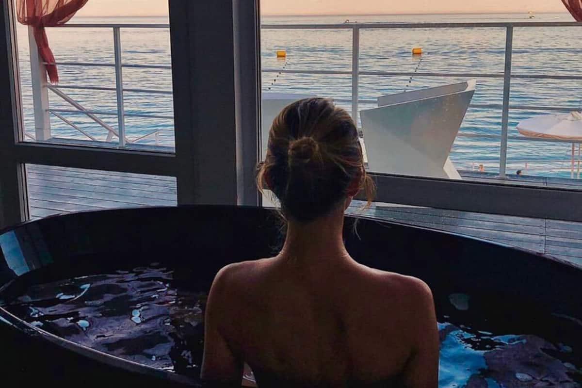 Eine Person liegt in einer Badewanne mit Blick auf das Meer durch große Fenster.