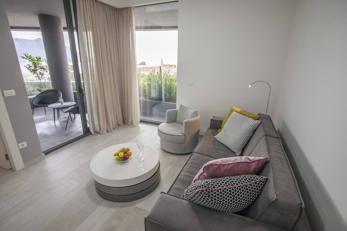 A modern living room with a grey sofa, a round coffee table with fruits and glasses, a patterned armchair, and a view of mountains through a large window.