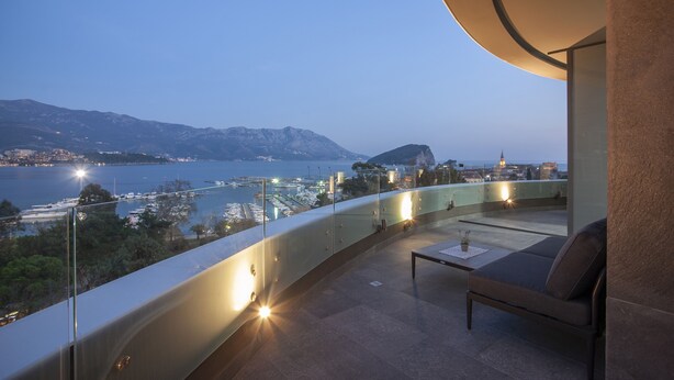A balcony with a curved glass railing overlooking a coastal cityscape at dusk. - Royal Gardens Paradise (Budva)