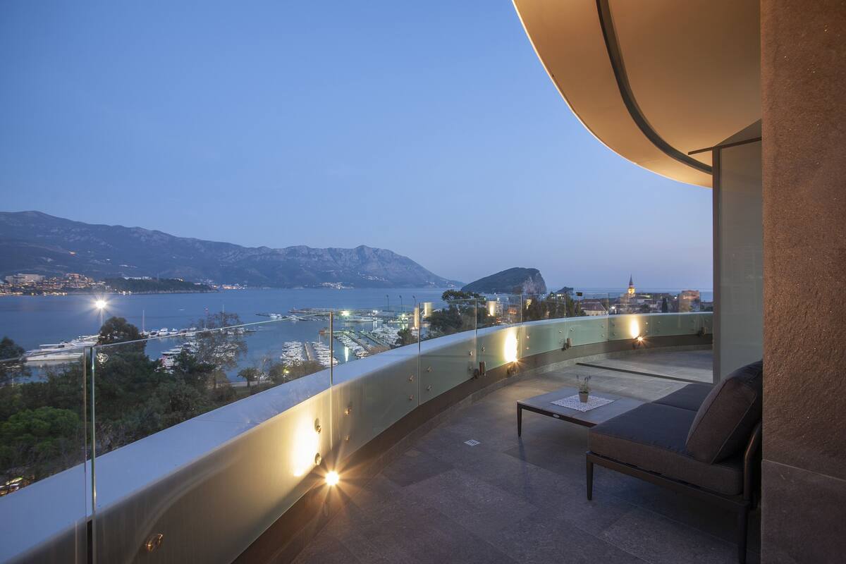 A balcony with a curved glass railing overlooking a coastal cityscape at dusk.