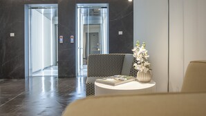 A modern lobby with a marble floor, two elevators, a patterned sofa, a small round table with a vase of flowers, and a tall floor lamp. - Royal Gardens Paradise (Budva)