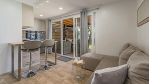 Un salon moderne comprenant un canapé, deux fauteuils, une petite table avec un livre ouvert, et un coin cuisine avec un réfrigérateur et un four.