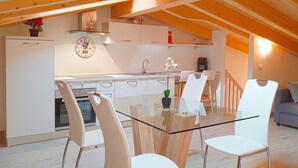 Una cocina moderna con armarios blancos, una mesa de comedor de vidrio y sillas blancas. Hay un sofá y una cafetera en la sala de estar.