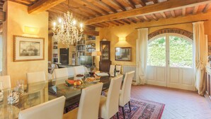 A dining area with a glass table, white chairs, a chandelier, bookshelves, and a large window with curtains. - Meriggio Barn (Capannori)