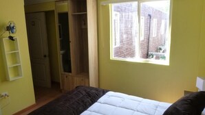 A bedroom with a bed, a window with a view of a brick building, a wardrobe, and a shelf with a fan. - Hotel Barrio Yungay (Santiago)