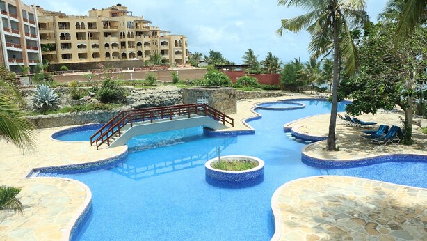 A resort pool area with multiple swimming pools, a bridge, and a palm tree. - Wild Flower Apartment (Mombasa)
