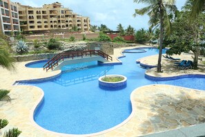 A resort pool area with multiple swimming pools, a bridge, and a palm tree. - Wild Flower Apartment (Mombasa)
