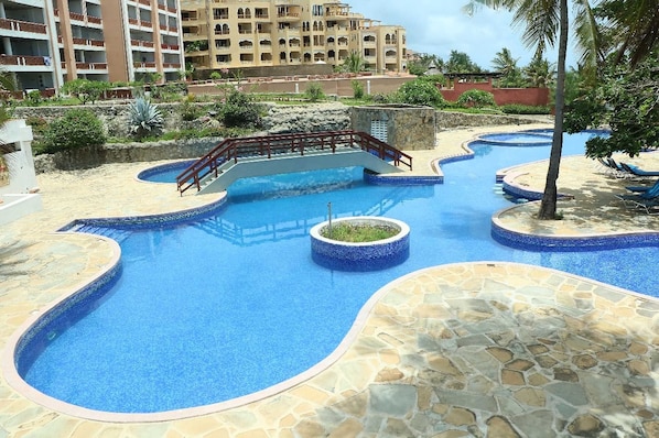 A swimming pool with a bridge, surrounded by stone tiles and palm trees.