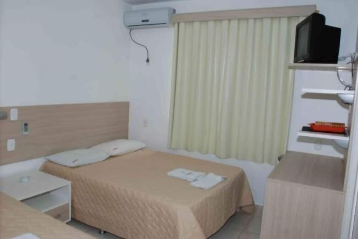 Quarto de hotel com cama, mesa de cabeceira, televisão e janela com cortinas.