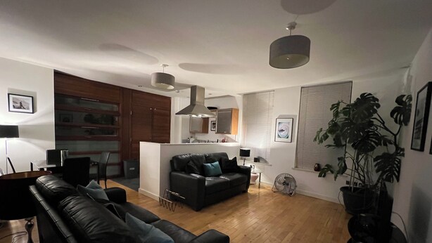 Un salon moderne avec un canapé en cuir noir, un coin repas avec une table en bois et une cuisine en arrière-plan.