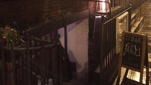 Une ruelle étroite, avec un bâtiment en briques sur la gauche et une structure en bois sur la droite. Une enseigne indiquant « CEDAR Gin & Tonic » est visible devant la structure en bois.