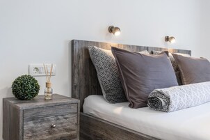 Cama bem arrumada com almofadas estampadas, uma pequena mesa de cabeceira com uma planta decorativa e luminárias de leitura fixadas na parede.