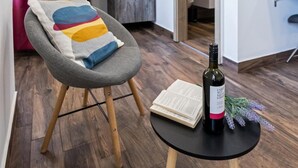 Ein modernes Wohnzimmer mit einem grauen Sessel, einem kleinen runden Tisch mit einer Weinflasche und einem Buch sowie einem Fernseher an der Wand.