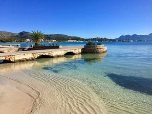 A clear waterfront with a sandy beach, a stone pier, and boats in the background. - 2 Bedroom Villa In Pollensa (Illes Balears)