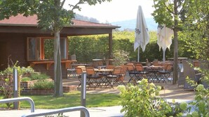 An outdoor patio area with tables and chairs, surrounded by greenery and a building with a sloped roof. - Landhotel Buchenkrug (Halle Westphalia)