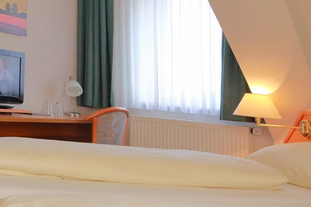 Hotellihuone, jossa on sänky, työpöytä, jossa on televisio, lamppu ja ikkuna, jossa on verhot.