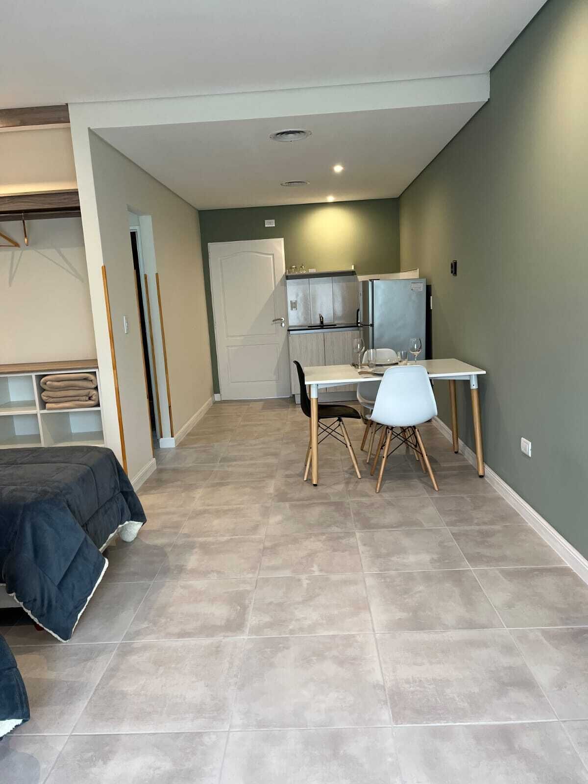 Quarto de hotel compacto com cama, pequena cozinha, área de refeições com mesa e cadeiras, e um guarda-roupa.