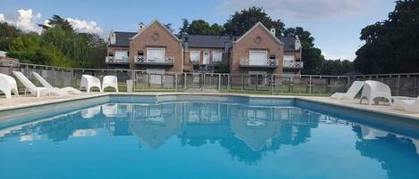 A swimming pool with white lounge chairs and a brick building with balconies in the background.