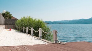 A wooden deck with a rope barrier, a sandy area with a red bucket, and a view of a lake with mountains in the background. - LA PLAYA: Villa with a Private Beach and All-Inclusive Sports  (Pettenasco)